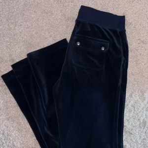 Mörkblå sammetsmjukisbyxor Juicy Couture - Mjukisbyxor i sammetsmaterial från Juicy Couture i färgen night sky. Byxorna har bred resår i midjan med snörning, fickor fram och bak med knappdetaljer samt broderad JC-logga framtill. Nyskick..
