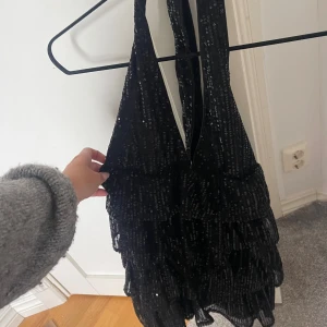 Svart paljettklänning med volanger - Supersnygg svart kortklänning med djup v-ringning och breda axelband. Klänningen är täckt av glittriga paljetter och har flera lager volanger och öppen i ryggen!