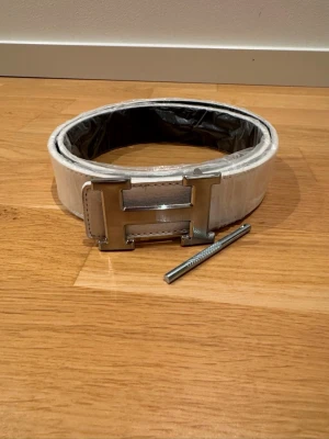 H-Bälte - Säljer ett stilrent vitt skärp med ett stort H-format metallspänne. Skärpet har en slät yta och tydliga sömmar längs kanterna. Passar perfekt för att lyfta vilken outfit som helst med en clean och modern look.