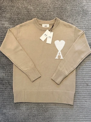 Beige stickad tröja från Ami - Säljer en beige stickad tröja från Ami Alexandre Mattiussi med ikoniskt vitt hjärta och A på bröstet. Tröjan har rund hals, ribbade muddar och nederkant samt lång ärm. Perfekt för dig som gillar stilrena och trendiga plagg.