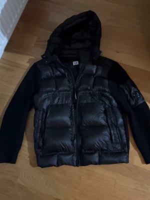 Svart dunjacka från C.P. Company - Svart dunjacka från C.P. Company med stickade ärmar och huva. Jackan har en quiltad front i glansigt material, dragkedja och en ikonisk linsdetalj på ärmen. Perfekt för kalla dagar och har en snygg, modern look. Är helst villig till att göra ett byte mot en monclear cardigan! 