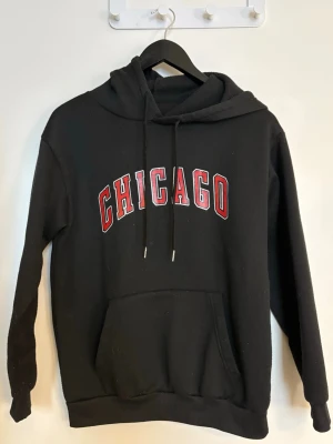 Dam svart hoodie med tryck på - Svart hoodie med trycket Chicago framtill i rött och vitt i Storlek XS. Tillverkad i 100 % polyester. Mjuk och bekväm med huva, snörning och magficka. 