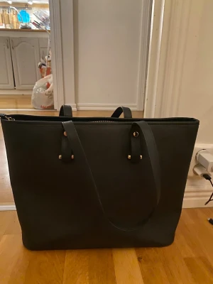 Svart axelväska från H&M - Stilren svart axelväska från H&M i syntetmaterial med guldfärgade detaljer och dragkedja upptill. Väskan har två långa handtag och rymligt innerfack med extra ficka och dragkedja. Perfekt för dig som vill ha plats för allt du behöver under dagen.