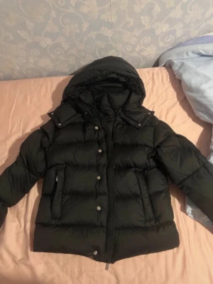 Svart pufferjacka med huva - Säljer en svart pufferjacka med huva och dragkedja framtill. Jackan har knappar längs öppningen och två stora fickor på sidorna. Perfekt för kalla dagar och riktigt skön att bära. Passar dig som gillar en enkel men snygg stil.