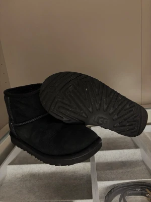Svarta UGG boots  - Säljer ett par klassiska svarta UGG boots i mjuk mocka med rund tå och platt sula. Insidan är fodrad för extra värme och komfort. Perfekta för kalla dagar och har en enkel, stilren look med UGG-logga bak på hälen.