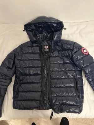 Mörkblå Canada Goose jacka - Snygg mörkblå dunjacka från Canada Goose med huva och klassisk logga på ärmen. Jackan har quiltad design, dragkedja framtill och två sidofickor med dragkedja. Perfekt för kalla dagar och enkel att packa ihop. Stilsäker och funktionell för dig som gillar streetwear.