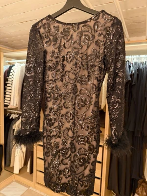 Svart glittrig kortklänning med fjädrar - Säljer en superfin svart kortklänning med glittriga paljetter och broderat mönster. Klänningen har långärmade ärmar med fjäderdetaljer vid ärmsluten och en djup v-ringning i ryggen. Normal i storlek o endast provad.
