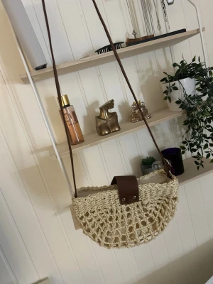 Beige axelväska i flätad bomull - Säljer en halvmåneformad axelväska i beige flätad bomull med brunt syntetband och snyggt lock. Väskan har ett öppet nätmönster och innerfack med dragkedja. Perfekt för dig som vill ha en chill och trendig accessoar till sommaren.
