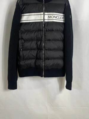 Svart moncler cardigan - Säljer en svart pufferjacka från Moncler med stickade ärmar och rygg. Jackan har vita ränder och Moncler-logga på bröstet samt dragkedja framtill med logodetalj. Perfekt för dig som vill ha en mix av sportig och stilren look.