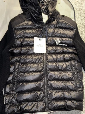Svart Moncler cardigan NY - Säljer en svart pufferjacka från Moncler med blank yta och stickade ärmar. Jackan har huva, dragkedja framtill och två fickor. Moncler-logga på bröstet och quiltad design på framsidan. Perfekt för dig som vill ha en stilren och exklusiv look.