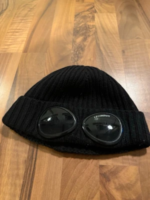 Svart mössa med goggles C.P. Company - Säljer en svart ribbstickad mössa från C.P. Company med ikoniska goggles framtill. Mössan är i mjukt material och har en bred uppvikt kant där glasögonen sitter fast. Perfekt accessoar för dig som vill sticka ut och hålla värmen.  Äkta såklart nypris runt 1400