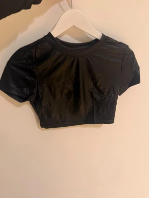 Svart croppad kortärmad topp - Snygg svart croppad topp med korta ärmar och rund halsringning. Tillverkad i ett glansigt syntetmaterial som ger en cool look. Perfekt att styla med höga jeans eller kjol.
