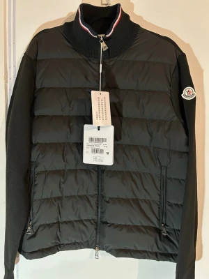 Svart pufferjacka  Moncler - Säljer en svart pufferjacka Moncler med stickade ärmar och rygg. Jackan har hög krage med röd, vit och blå rand, två dragkedjefickor och hel dragkedja framtill. Klassisk Moncler-logga på ärmen och quiltad front i syntetmaterial.