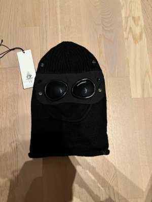 Svart balaclava från C.P. Company - Svart stickad balaclava från C.P. Company med ikoniska goggles och detaljer i metall. Snygg och funktionell för kalla dagar, perfekt för dig som vill sticka ut. Märkeslapp på kanten och justerbar öppning för ansiktet.