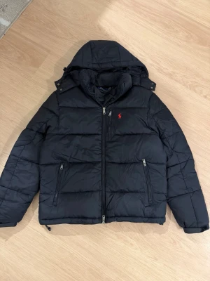 Svart pufferjacka från Polo Ralph Lauren - Säljer en svart pufferjacka från Polo Ralph Lauren med huva och dragkedja framtill. Jackan har två sidofickor med dragkedja och den klassiska röda loggan broderad på bröstet. Perfekt för kalla dagar och riktigt snygg till streetwear-stilen. I have the receipt for it too