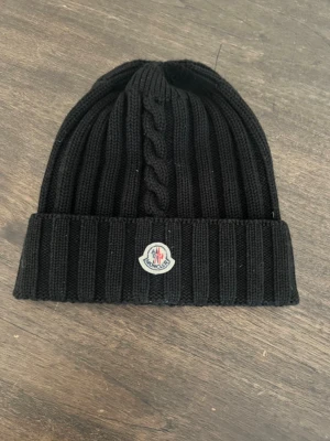 Svart stickad mössa från Moncler - Säljer en svart ribbstickad mössa från Moncler med klassisk logga framtill. Mössan har en bred uppvikt kant och ett snyggt flätmönster mitt fram. Perfekt accessoar för kyliga dagar och riktigt stilren till alla outfits.