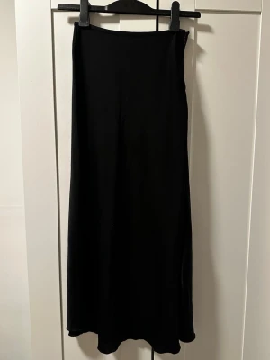 Svart satinkjol från Amisu - Säljer en svart lång satin kjol från Amisu i storlek 36. Kjolen har en enkel och stilren design med lätt utsvängd form och dragkedja i sidan. Materialet känns mjukt och följsamt, perfekt för dig som gillar minimalistisk stil.