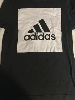 Svart Adidas t-shirt med stor logga - Svart t-shirt från Adidas med stor vit fyrkant och svart Adidas-logga på bröstet. Klassisk rund halsringning och korta ärmar. Tillverkad i mjuk bomull som känns skön mot huden. Perfekt för dig som gillar sportig och enkel stil.