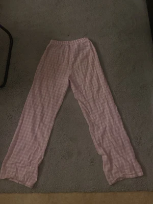 Rosa rutiga byxor från Gina Tricot - Säljer ett par mjuka och sköna byxor från Gina Tricot i storlek 158/164. Byxorna är ljusrosa med vitt rutmönster, har resår i midjan och snörning för extra komfort. Perfekta för chill dagar hemma.