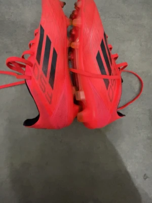Röda Adidas F50 fotbollsskor - Säljer ett par röda Adidas F50 fotbollsskor med svarta detaljer och klassiska tre ränder på sidan. Skorna har snörning och är tillverkade i syntetmaterial för lätt känsla på planen. Perfekta för dig som vill ha speed och kontroll på gräs.