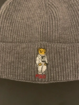Grå stickad mössa från Polo Ralph Lauren - Säljer en grå ribbstickad mössa från Polo Ralph Lauren med broderad nalle i astronautdräkt framtill. Klassisk design med uppvikt kant och mjukt material som håller dig varm under kyliga dagar.