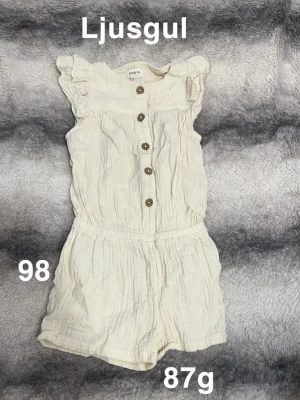 Ljusgul byxdress med knappar - Supersöt ljusgul byxdress från Newbie med träknappar framtill och volangdetaljer på axlarna. resår i midjan för skön passform. Perfekt för varma dagar!🌸 Ljusgul. Fint använt skick. Storlek 98