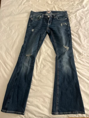 Bootcut jeans från Gina Tricot - Mörkblå bootcut jeans från Gina Tricot med slitna detaljer och kontrastsömmar. Klassisk femficksmodell med broderade bakfickor och låg midja. Tillverkade i jeansmaterial med dragkedja och knapp framtill under linningen.