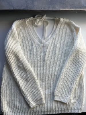 Vit stickad tröja med knyt i ryggen - Säljer en krämvit stickad tröja med v-ringning och snygg knytning i ryggen. Tröjan har lång ärm och är ribbstickad för en mysig och trendig look. Perfekt för lager på lager under höst och vinter.