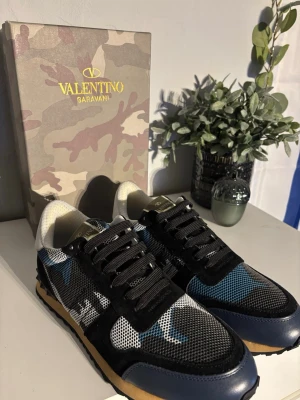 Valentino Garavani Rockrunner sneakers - Säljer ett par Valentino Garavani Rockrunner sneakers med blå, svart och vit camouflagemönstrad ovandel i mesh och mocka. Skorna har svarta snören, gummisula i beige och svart samt detaljer i skinn. Ikonisk logga på plösen och undersidan.