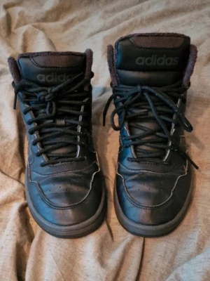 Svarta Adidas sneakers med foder - Säljer ett par svarta Adidas sneakers med högt skaft och mjukt foder. Skorna har klassiska tre ränder på sidan, snörning och rund tå. Insidan är fodrad för extra värme. Perfekta för kyligare dagar. Dom är välanvända men har fortfarande lite kvar att ge. Perfekt för växande fötter som inte kan ha skor länge än en säsong. De är noppriga och sulan har släppt lite vid tårna på högerfoten. Stl 39½