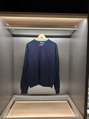 Mörkblå ulltröja från Polo Ralph Lauren - Klassisk mörkblå stickad tröja från Polo Ralph Lauren i 100% lammull. V-ringad modell med ribbade muddar och ikoniska broderade loggan på bröstet. Perfekt för dig som gillar stilrena och tidlösa plagg.                                                                  Längd:63cm.     Bredd:42cm