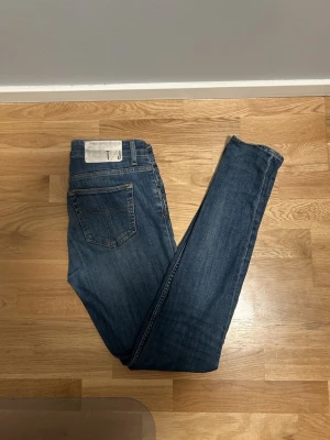 Blå jeans från Tiger of Sweden - 29/34 - Säljer ett par klassiska blåa skinnyjeans från Tiger of Sweden. De har en snygg tvätt, fem fickor och guldfärgade sömmar. Passformen är smal med raka ben och normal midja. 