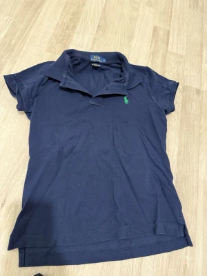 Mörkblå piké från Polo Ralph Lauren - Klassisk mörkblå pikétröja från Polo Ralph Lauren med korta ärmar och krage. Tröjan har den ikoniska gröna loggan broderad på bröstet och är tillverkad i mjuk bomull. Perfekt till jeans eller shorts för en clean och enkel stil.
