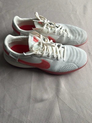 Nike Gato vita och röda sneakers - Säljer ett par Nike Gato Futsal skor sneakers i vitt och rött med klassisk swoosh på sidan. Skorna har snörning, perforerade detaljer på tån och ovandel i mocka och syntet. Snygg kontrast mellan den vita basen och de röda detaljerna. Perfekta för dig som gillar sportig stil.