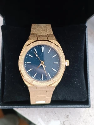 Guldklocka – "Frosted" Finish & Galaxy-urtavla ✨ - Säljer en klocka som har en unik "frosted"  textur på både boett och länk.                                  Design: Klassisk oktagonal boett med integrerad länk. Urtavla: Mörkblå/svart tavla med "stardust"-effekt som påminner om en stjärnhimmel. Skick: Helt ny och oanvänd (skyddsplasten sitter kvar på baksidan/länken). Stil: Perfekt för dig som gillar "Iced out"-looken men vill ha något mer stilrent.