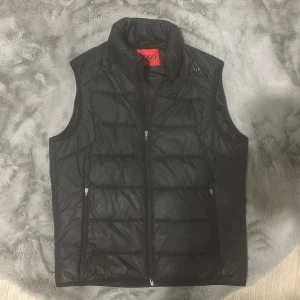 Svart dunväst från Hugo Boss - Snygg svart dunväst från Hugo Boss med diskret logga på bröstet och hög krage. Västen har två fickor med dragkedja och quiltad design. Perfekt att slänga över en hoodie eller tröja för en clean look.