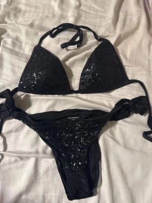Svart paljettbikini från Calzedonia - Säljer en svart bikini från Calzedonia med glittriga paljetter på både överdel och underdel. Bikinin har knyt i nacken och på sidorna för justerbar passform. Perfekt för dig som vill sticka ut på stranden!