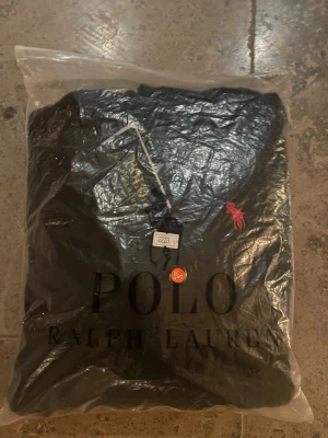 Svart kabelstickad tröja Polo Ralph Lauren - Helt ny, aldrig använd, kabelstickad, svart tröja från Polo Ralph Lauren med klassisk röd logga broderad på bröstet. Tröjan har rund halsringning och långa ärmar. Perfekt för dig som vill ha en stilren och tidlös look med premiumkänsla.