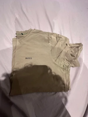 Beige BOSS t-shirt med tryck - Säljer en beige t-shirt från BOSS i storlek XL. T-shirten har rund hals, korta ärmar och ett stilrent svart BOSS-tryck på bröstet. Materialet är mjuk bomull och modellen har en normal passform. Perfekt för dig som gillar enkel och clean stil.