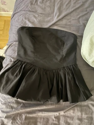 Svart volang tubtopp - Säljer en svart axelbandslös tubtopp med bred volang nedtill. Toppen har en figurnära passform upptill och är sydd i ett mjukt tyg som ger en snygg siluett. Perfekt för dig som vill ha en statement piece till garderoben.