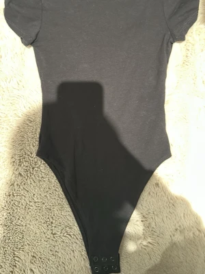 Svart basic body med kort ärm XXS från H&M - En enkel svart body från H&M med korta ärmar och rundad urringning. Modellen har tryckknappar i grenen för enkel på- och avtagning. Perfekt att styla med jeans eller kjol för en clean look. Har aldrig använt utan bara legat i garderoben och fick hänga med ut ur min rensning.