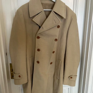 Beige rock från Dolce & Gabbana i riktigt bra skick - Säljer en klassisk beige vintage rock från Dolce & Gabbana med dubbelknäppning och stora slag. Jackan har snygga bruna knappar, två fickor fram och detaljer vid ärmsluten. Fodret är mönstrat med D&G-logga för en exklusiv känsla. Perfekt för dig som vill ha en stilren och tidlös look. Otroligt bra skick!