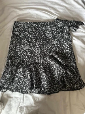 Svartvit prickig omlottkjol - Säljer en svartvit kort omlottkjol med prickigt mönster och volangkant. Kjolen har en knytning i midjan som ger en snygg detalj och sitter skönt på kroppen. Perfekt för dig som gillar trendiga och lekfulla plagg.