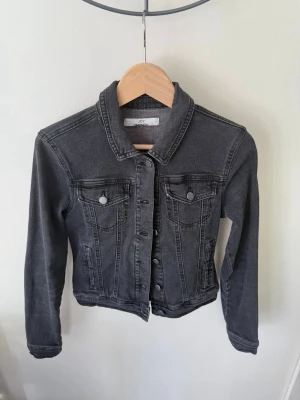 Mörkgrå jeansjacka från JDY - Snygg mörkgrå jeansjacka från JDY med klassisk krage, knappar framtill och två bröstfickor. Jackan har lång ärm och en croppad passform som ger en cool vibe till din outfit.