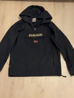 Mörkblå vindjacka från Napapijri - Säljer en mörkblå vindjacka från Napapijri med huva och stor ficka framtill. Jackan har dragkedja upptill, broderad Napapijri-logga och norsk flagga på bröstet. Tillverkad i slitstark polyester och har en loose passform. Perfekt för dig som vill ha en stilren och funktionell jacka.