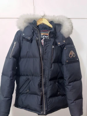 Svart Moose Knuckles pufferjacka L - Säljer en mörk blå pufferjacka från Moose Knuckles med stor fluffig päls runt huvan, dragkedjor på ärmar och fram, samt snygga metallknappar. Jackan har flera fickor och ikonisk logga på ärmen. Perfekt för kalla dagar och riktigt stilren. Ej använd! Köpt på Urban Modern, Kvitto finns.