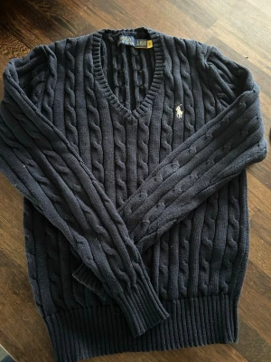 Mörkblå kabelstickad tröja Polo Ralph Lauren - Klassisk mörkblå kabelstickad tröja från Polo Ralph Lauren med v-ringning och broderad logga på bröstet. Tröjan har långa ärmar och ribbade muddar vid ärmslut och nederkant. Perfekt för dig som gillar tidlös och preppy stil. Använd ett par gånger, köpt ny för 1500 kr
