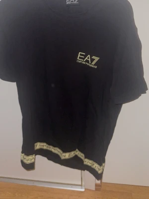Svart EA7 Emporio Armani t-shirt - Svart t-shirt från EA7 Emporio Armani med gul logga på bröstet och gul textdetalj längs nederkanten. Klassisk rund hals och korta ärmar. Perfekt för dig som gillar stilrena och exklusiva plagg.
