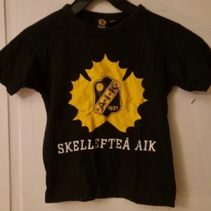 Svart Skellefteå AIK t-shirt barn - Cool svart t-shirt från Skellefteå AIK Hockey med stort gult klubbmärke och vit text på bröstet. Klassisk rund hals och korta ärmar. Perfekt för unga fans som vill visa sitt stöd för laget. Materialet är mjuk bomull. Hund finns i hemmet. Kolla in mina andra annonser för samfrakt! 