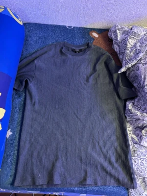 Mörkgrå t-shirt från SMOG XS - En mörkgrå t-shirt från SMOG i relaxed fit. T-shirten har korta ärmar, rund halsringning och är tillverkad i ett strukturerat polyestertyg som ger en cool känsla. Perfekt för dig som gillar enkel och stilren design.